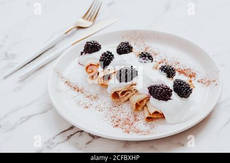 Deliziose crepes con yogurt e more servite sul piatto con cannella vicino argenteria su tavolo di marmo Foto Stock