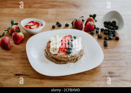 Una pila di gustosi pancake serviti sul piatto con pezzi di banana e fragola e mirtilli freschi con fiocchi di cocco durante colazione Foto Stock