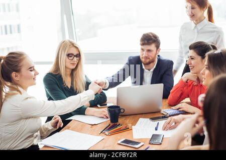 Benvenuto a bordo! Due bei uomini che scuotono le mani con un sorriso mentre si siede sul divano in ufficio con i loro colleghi Foto Stock