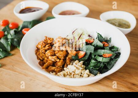 pezzi di pollo arrosto yummy messi sul piatto vicino alla rucola e insalata di pomodoro con cavolo tritato e grano su legno tavolo vicino salsa e argenteria Foto Stock