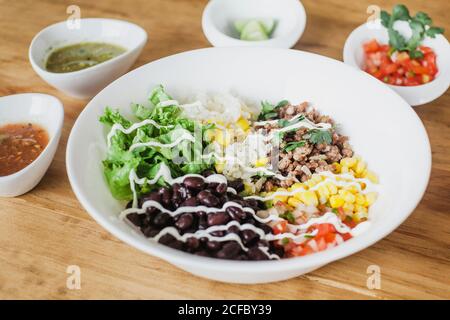 lattuga e fagioli con pomodori tritati messi vicino a chicchi di mais e grano in ciotola su tavola di legno vicino varie salse con calce e argenteria Foto Stock