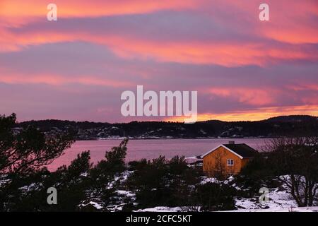 Vivace cielo rosa tramonto su cottage rurale circondato da alberi sulla sponda remota del fiume innevato Foto Stock
