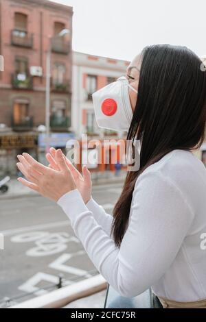 Vista laterale di una giovane femmina positiva in posizione eretta con maschera respiratore sul balcone e applauding medici specialisti durante l'epidemia di coronavirus Foto Stock