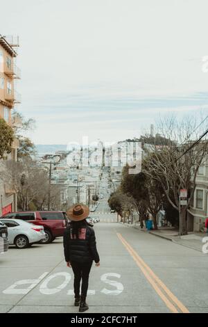 Vista posteriore di una donna irriconoscibile viaggiatore in abiti alla moda e. cappello in piedi vicino allo stop sulla strada che conduce verso la collina Durante la passeggiata e la visita turistica della città di San Francisco nel nuvoloso giorno Foto Stock