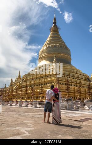 Vista posteriore della romantica giovane coppia che trascorre la luna di miele in antico Città di Bagan in Myanmar e ammirando la splendida architettura mentre si è in piedi e abbracciando di fronte alla pagoda dorata in giornata di sole Foto Stock