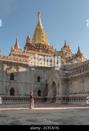 Vista laterale di Donna in abiti tradizionali godendo di architettura antica, mentre in piedi nel cortile di fronte al tempio buddista in pietra a Bagan Myanmar in giorno di sole con cielo blu Foto Stock