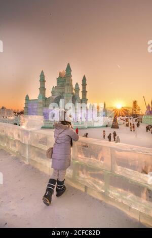 Vista posteriore di donna anonima in outwear caldo che pende sopra barriera di ghiaccio e ammirare sculture di neve in serata durante l'inverno festival Foto Stock