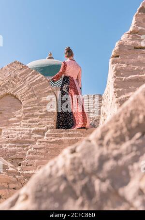 Da sotto vista posteriore di femmina calmo in lungo tradizionale vestitevi in piedi su scale di pietra e guardate lontano contro il vecchio Cupola blu del mausoleo di Pahlavan Mahmud a Khiva Foto Stock