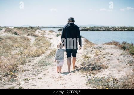 Vista posteriore del giovane uomo in abiti scuri che tengono le mani con il bambino e camminando intorno spiaggia sabbiosa con verde asciutto in giornata di sole Foto Stock