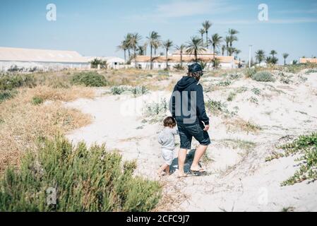 Vista posteriore del giovane uomo in abiti scuri che tengono le mani con il bambino e camminando intorno duna con verde e palma in giornata di sole Foto Stock