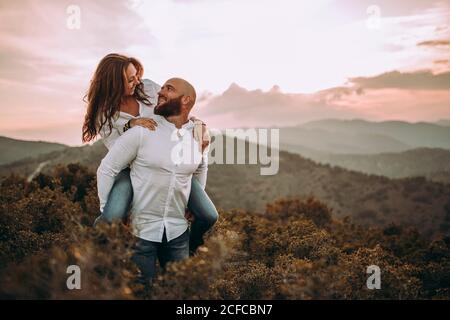 Coppia amorevole in camicia bianca corrispondente che si diverte e cavalcare piggyback a pendio di montagna erboso Foto Stock