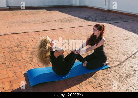 Vista laterale ad angolo alto della donna matura con una bambina che fa asana seduto mentre pratica yoga partner insieme sulla terrazza soleggiata sul tetto Foto Stock
