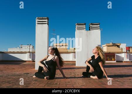 Vista laterale della donna di mezza età con la figlia giovane seduta in Ardha Matsyendrasana variazione posa mentre pratica yoga insieme sulla terrazza sul tetto in giornata di sole in città Foto Stock