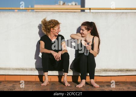Giovane donna allegra con madre di mezza età in abbigliamento sportivo che parla e ride mentre si siede con una bottiglia d'acqua sul tetto e riposarsi dopo la pratica di yoga insieme Foto Stock