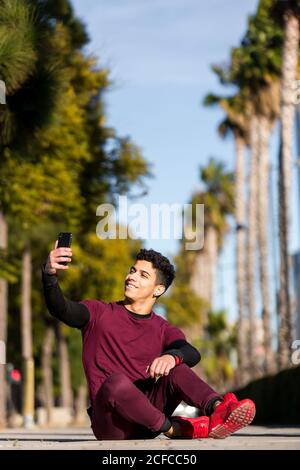 Giovane ragazzo etnico allegro in sportswear sorridente e prendendo selfie mentre si siede sul percorso durante l'allenamento fitness in giornata di sole in città Foto Stock