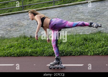 Vista laterale della giovane femmina in abbigliamento sportivo e lame a rulli equilibrio sulla gamba mentre divertente durante il fine settimana Foto Stock