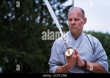 Determinato maschio di mezza età in kimono focalizzare e usando la spada mentre si allenano da soli in giardino Foto Stock