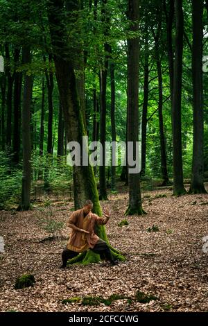 Ragazzo asiatico in marrone uniforme gesticulating mentre fa arti marziali esercizio fisico durante l'allenamento nella foresta verde Foto Stock