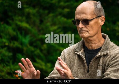 Calvo uomo anziano in occhiali da sole che eseguono l'esercizio durante le arti marziali formazione su sfondo sfocato di alberi verdi in giardino Foto Stock