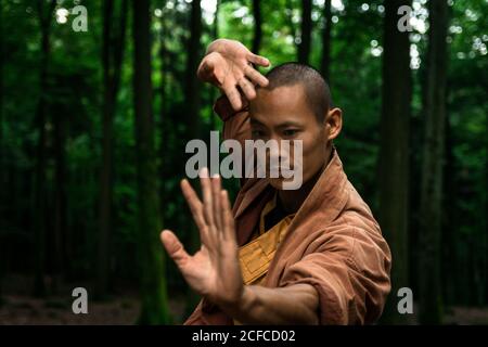 Ragazzo asiatico in marrone uniforme gesticulating mentre fa arti marziali esercizio fisico durante l'allenamento nella foresta verde Foto Stock