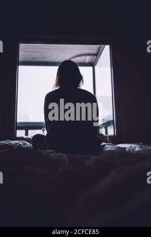 Dalla silhouette del soffietto di anonima Donna solitaria seduta sul letto contro la finestra nella stanza scura a casa Foto Stock