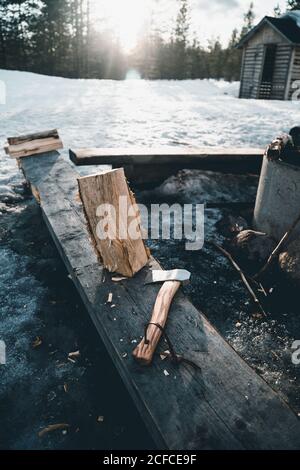 Falò bruciante e tronco con ascia collocato vicino a piccola cabina di lumberjack in foresta innevata nella giornata invernale in campagna Della Finlandia Foto Stock