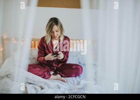 Giovane donna graziosa in pigiama seduto sul letto guardando lo smartphone Foto Stock