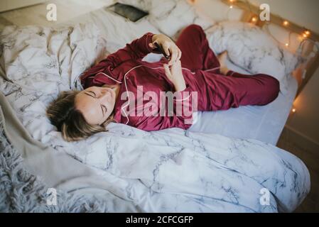 Giovane donna graziosa in pigiama sdraiata sul letto guardando lo smartphone Foto Stock