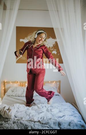 Giovane donna sorridente felice in pigiama con cuffie che ballano a letto al mattino Foto Stock