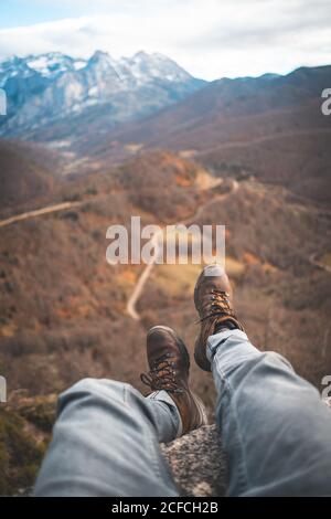 Il viaggiatore di raccolto in stivali marroni seduti con le gambe penzolanti bordo sulla roccia e godendo di vista mozzafiato della foresta e. strada Foto Stock
