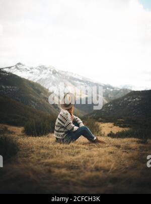 Vista laterale di irriconoscibile donna affascinante pensivo con capelli rossi in caldo maglione seduto sul prato in montagna in giorno grigio Foto Stock