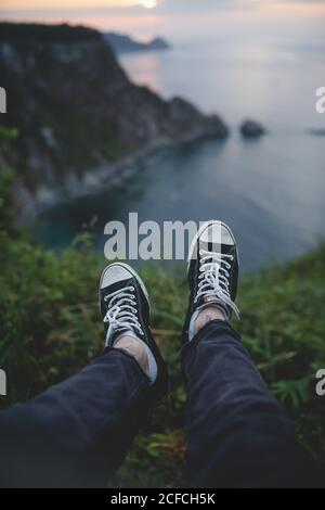 Da sopra raccolto piedi estenati in sneakers di uomo anonimo seduto sul bordo della collina sullo sfondo della scenografica stagione serale delle Asturie, Spagna Foto Stock