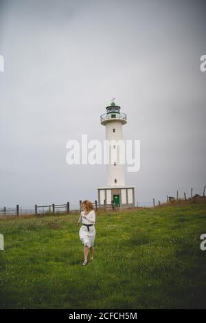 Spaventato rosso Donna in vestito bianco che corre pensieroso in campo contro faro in tempo nuvoloso nelle Asturie, Spagna Foto Stock