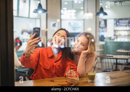 Due giovani amici della donna con tazze di caffè fresco sorridenti e in posa per selfie mentre si siede al tavolo in un accogliente ristorante con una bevanda Foto Stock