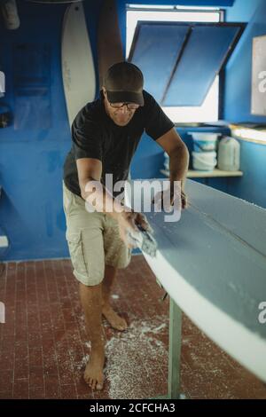 Woodworker che scuoiano diligentemente tavole da surf in officina Foto Stock