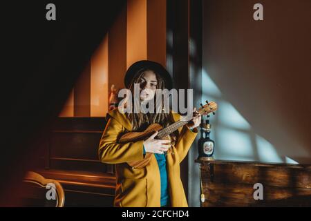 Hipster millennial femmina con dreadlock indossando cappotto giallo e nero cappello che suona la chitarra hawaiana ukulele mentre si trova in una vintage scura camera Foto Stock