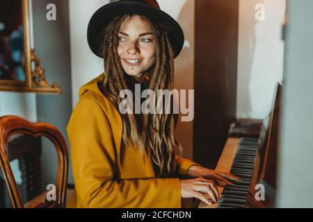 Vista laterale della femmina hipster felice con i dreadlock che indossa giallo cappotto e cappello nero suonano pianoforte mentre si siede in retrò camera in stile Foto Stock