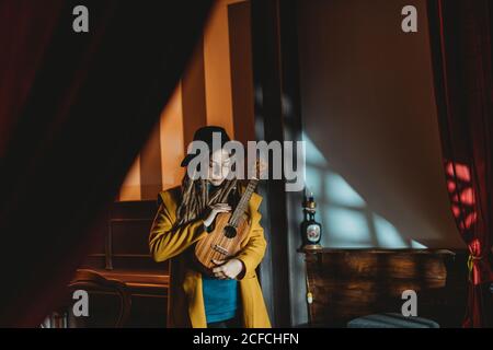 Hipster millennial femmina con dreadlock indossando cappotto giallo e nero cappello che suona la chitarra hawaiana ukulele mentre si trova in una vintage scura camera Foto Stock