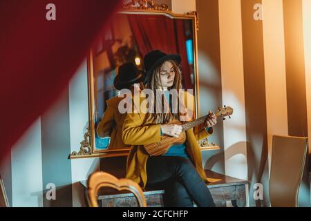 Giovane donna elegante con Dreadlocks indossando cappotto giallo e cappello nero seduto sul vecchio tavolo di legno indietro a specchio e suonando ukulele chitarra hawaiana in camera con mobili antichi Foto Stock