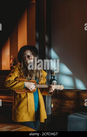 Hipster millennial femmina con dreadlock indossando cappotto giallo e nero cappello che suona la chitarra hawaiana ukulele mentre si trova in una vintage scura camera Foto Stock