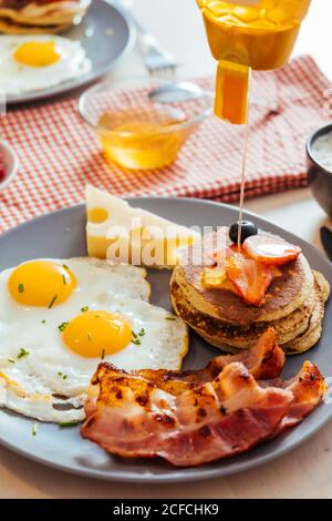 Dall'alto del piatto servito con uova fresche fritte con formaggio e pancetta e pancake con frutti di bosco e miele sul tavolo per la colazione Foto Stock