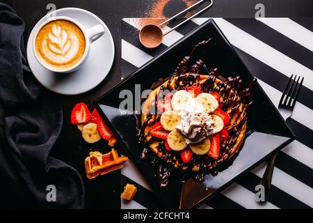 Vista dall'alto del cappuccino in tazza bianca sul tavolo con piatto di cialda rotonda con banana e fragola con salsa al cioccolato e panna montata Foto Stock