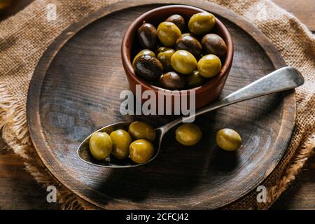 Da sopra piatto di legno ciotola e cucchiaio con nero e. olive verdi poste su tessuto rustico su tavola di legno Foto Stock