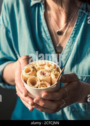 Gelato arrotolato in tazza conica in mani Donna. Donna in jeans camicia contiene cono tazza con vaniglia stile tailandese laminato gelato. Verticale Foto Stock