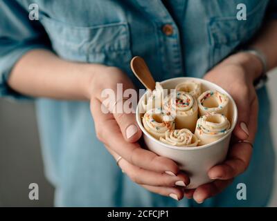 Gelato arrotolato in tazza conica in mani Donna. Donna in jeans camicia contiene cono tazza con vaniglia stile tailandese laminato gelato Foto Stock