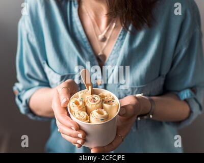 Gelato arrotolato in tazza conica in mani Donna. Donna in jeans camicia contiene cono tazza con vaniglia stile tailandese laminato gelato Foto Stock