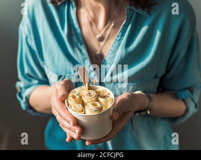 Gelato arrotolato in tazza conica in mani Donna. Donna in jeans camicia contiene cono tazza con vaniglia stile tailandese laminato gelato Foto Stock