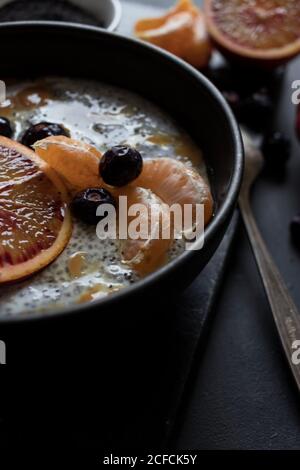 Dall'alto semi di chia pudding con fette di arancia, mandarini e olive in ciotola vicino al cucchiaio e pompelmo su tavola scura Foto Stock