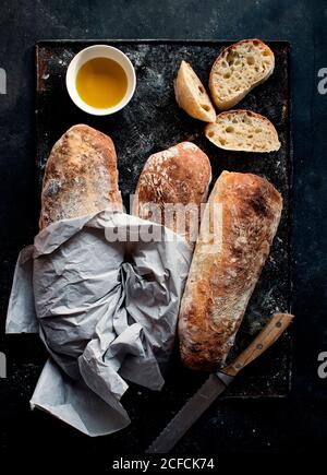 Pane ciabatta su tavola rustica vicino all'olio d'oliva e coltello con fette di pane su sfondo scuro Foto Stock