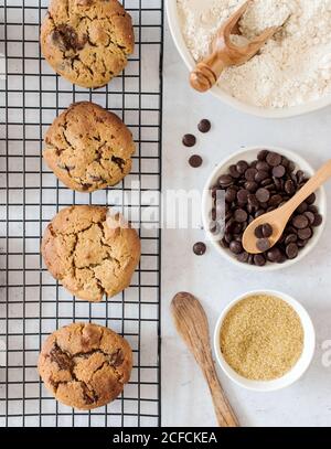 Vista dall'alto di deliziosi biscotti fatti in casa appena sfornati con cioccolato patatine poste sulla griglia della cucina vicino alle ciotole con ingredienti Foto Stock